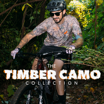 A man wearing a Timber Camo collection cycling jersey and shorts rides a mountain bike through dense green forest. The text 'THE TIMBER CAMO COLLECTION' is overlaid on the left side.