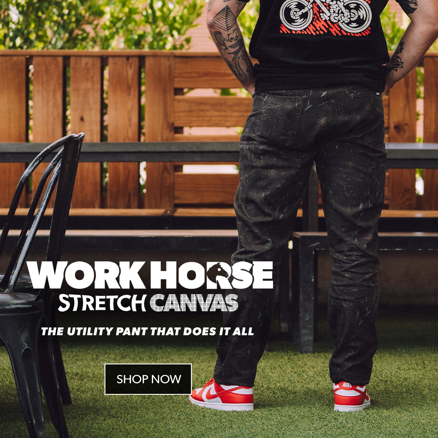 A person standing outdoors in front of a wooden bench and green grass, wearing dark work pants with white paint splatters and a black graphic T-shirt. Large text reads 'WORK HORSE STRETCH CANVAS' with a 'SHOP NOW' button below.