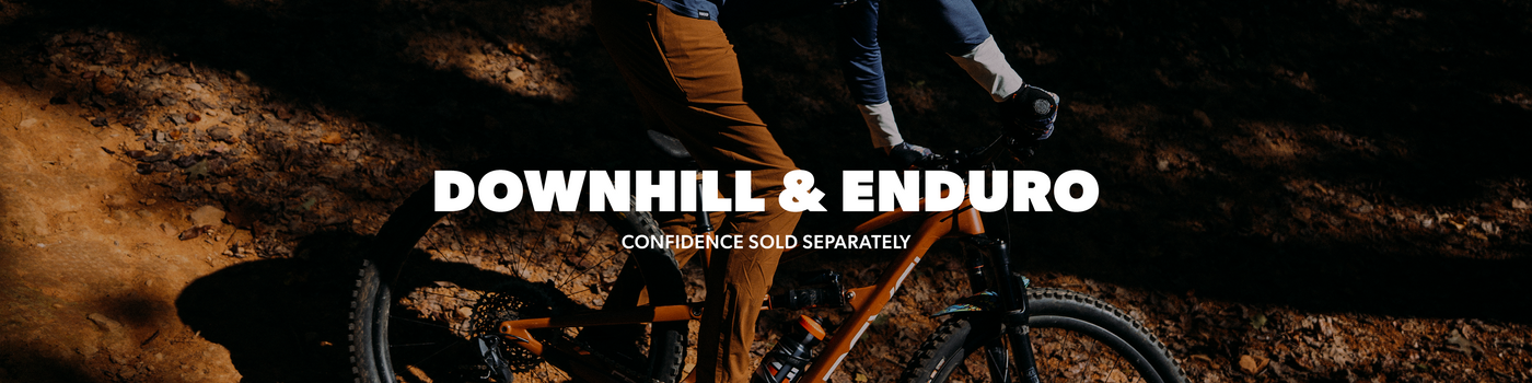 A person riding a mountain bike on a dirt trail at night, with large white text reading 'DOWNHILL & ENDURO' and smaller text 'CONFIDENCE SOLD SEPARATELY' across the center.