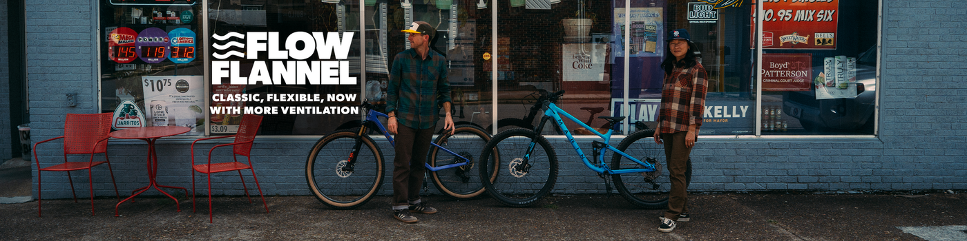 Two people standing with mountain bikes in front of a storefront with the text 'FLOW FLANNEL' overlaid on the left. The scene is outdoors on a street or sidewalk, with a blue wall, shop windows, red chairs, and two bikes visible: one dark blue and one bright blue.