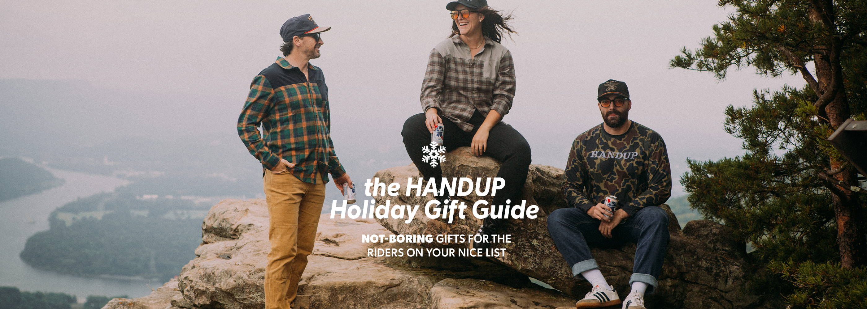 Three people sitting on a cliff overlooking a scenic landscape with 'The HANDUP Holiday Gift Guide' text overlay.