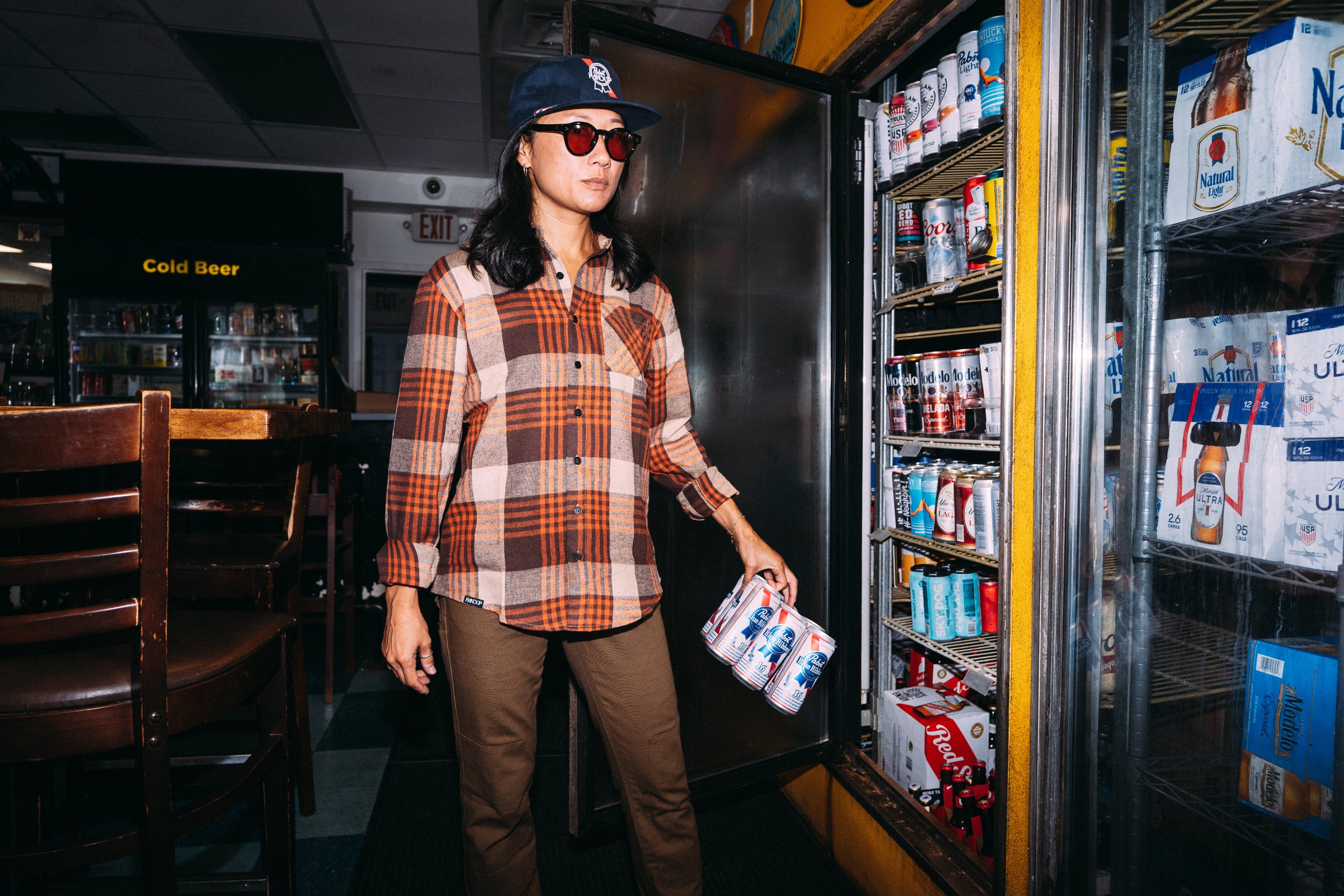 A woman wearing a brown and orange plaid flannel shirt, brown pants, a navy cap, and sunglasses stands next to an open refrigerator in a convenience store, holding a six-pack of bottled beverages.
