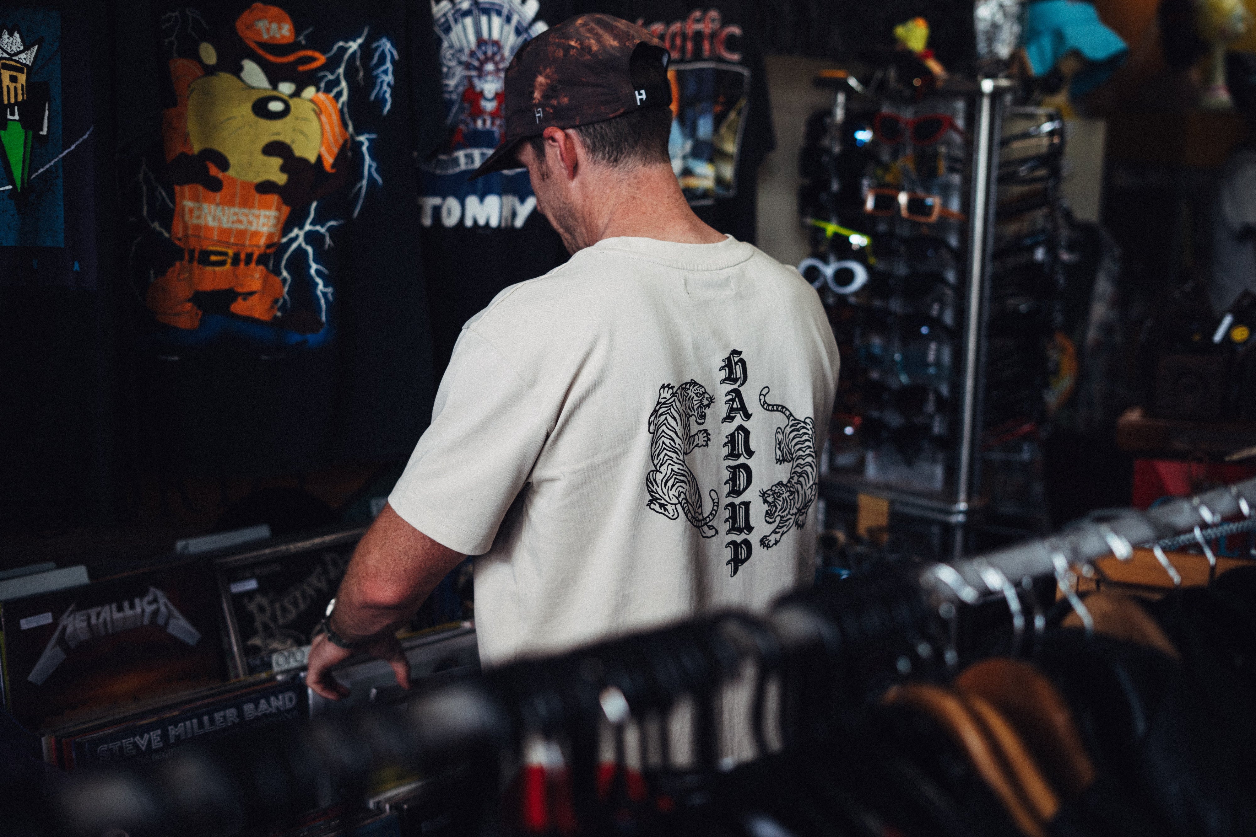 A man viewed from behind wearing a cream-colored t-shirt with a black graphic print on the back, standing inside a retail store.