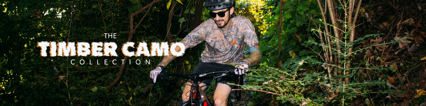 A man wearing a Timber Camo collection cycling jersey and shorts rides a mountain bike through dense green forest. The text 'THE TIMBER CAMO COLLECTION' is overlaid on the left side.