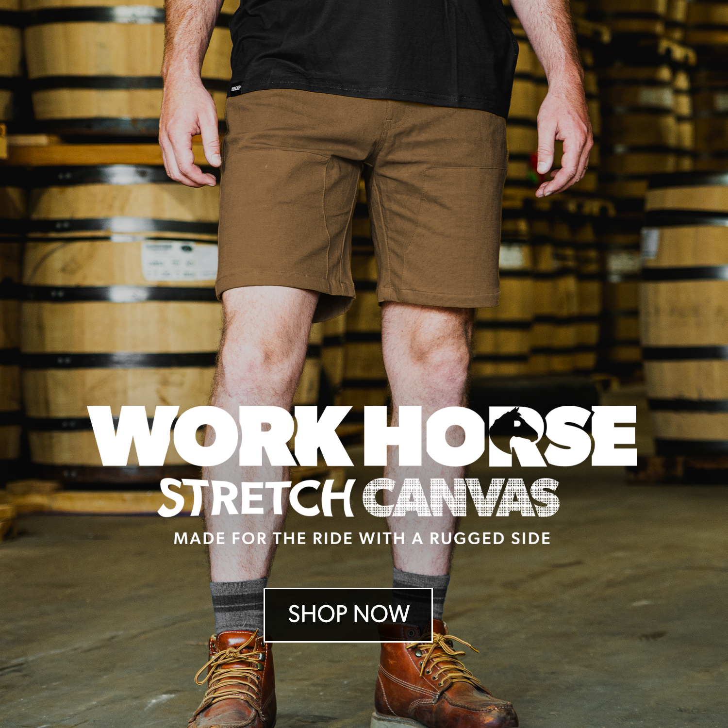 A person wearing brown stretch canvas work shorts, a black t-shirt, gray socks, and brown work boots stands in front of a blurred background of wooden barrels. Large text overlays read 'WORK HORSE STRETCH CANVAS' with a 'SHOP NOW' button below.