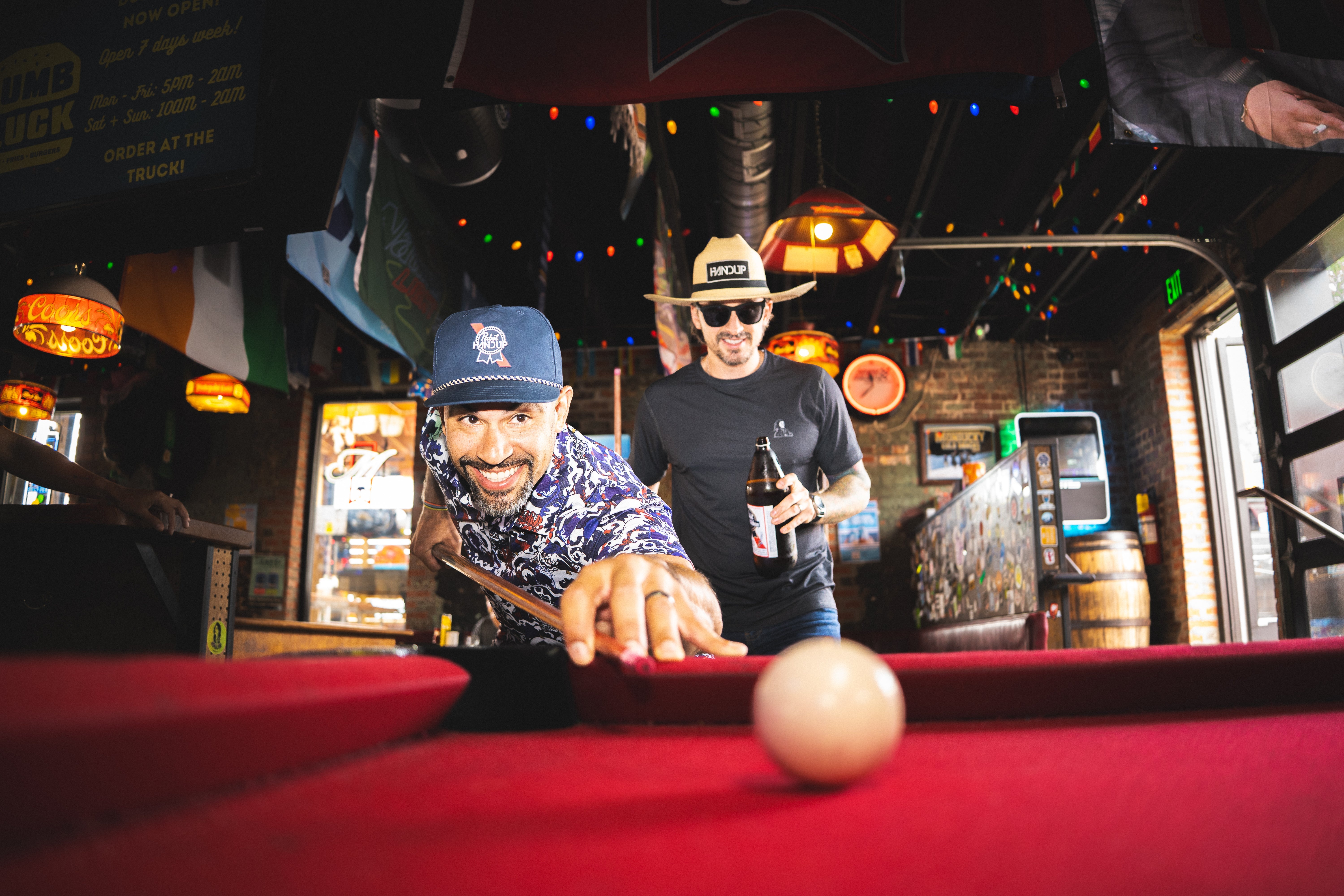 two men playing pool in a bar setting wearing hats