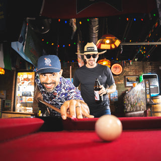 two men playing pool in a bar setting wearing hats