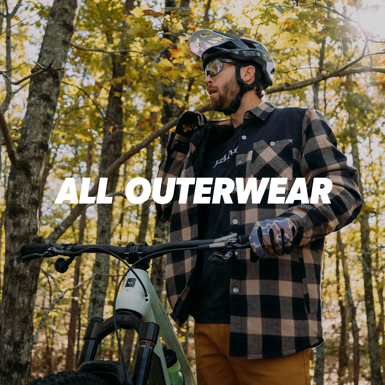 A man adjusting a plaid jacket wearing a helmet and holding a mountain bike in a forest setting 