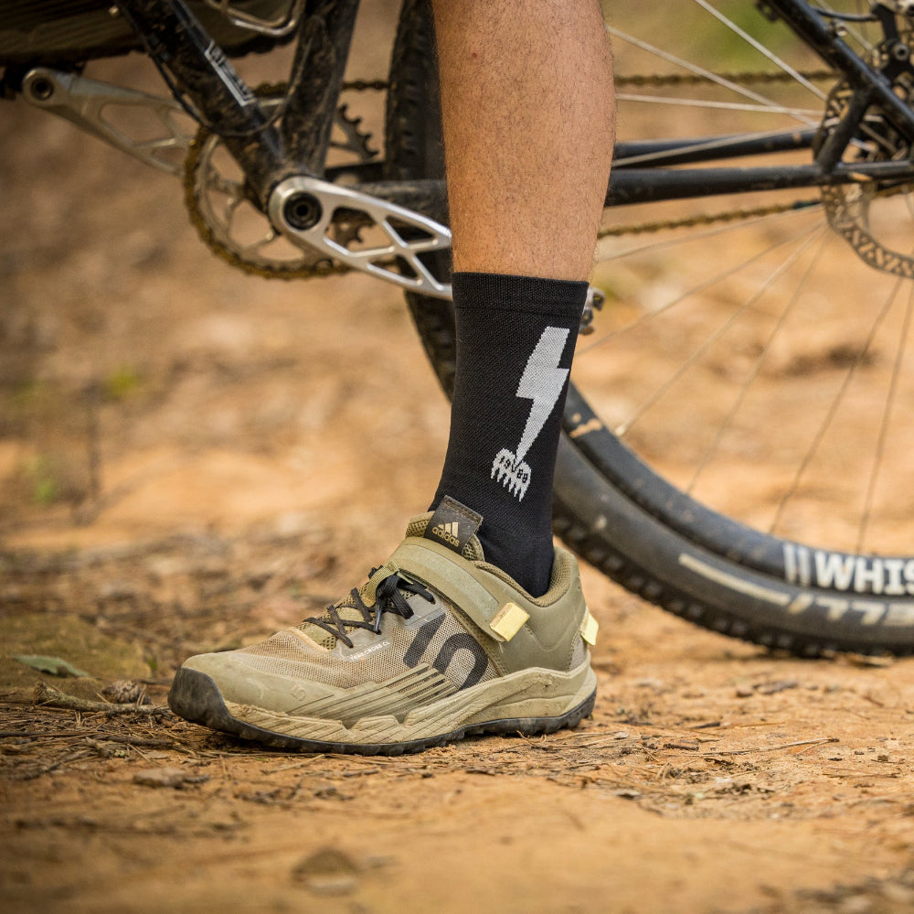 Person wearing cycling shoes and socks with a bike in the background on a dirt path