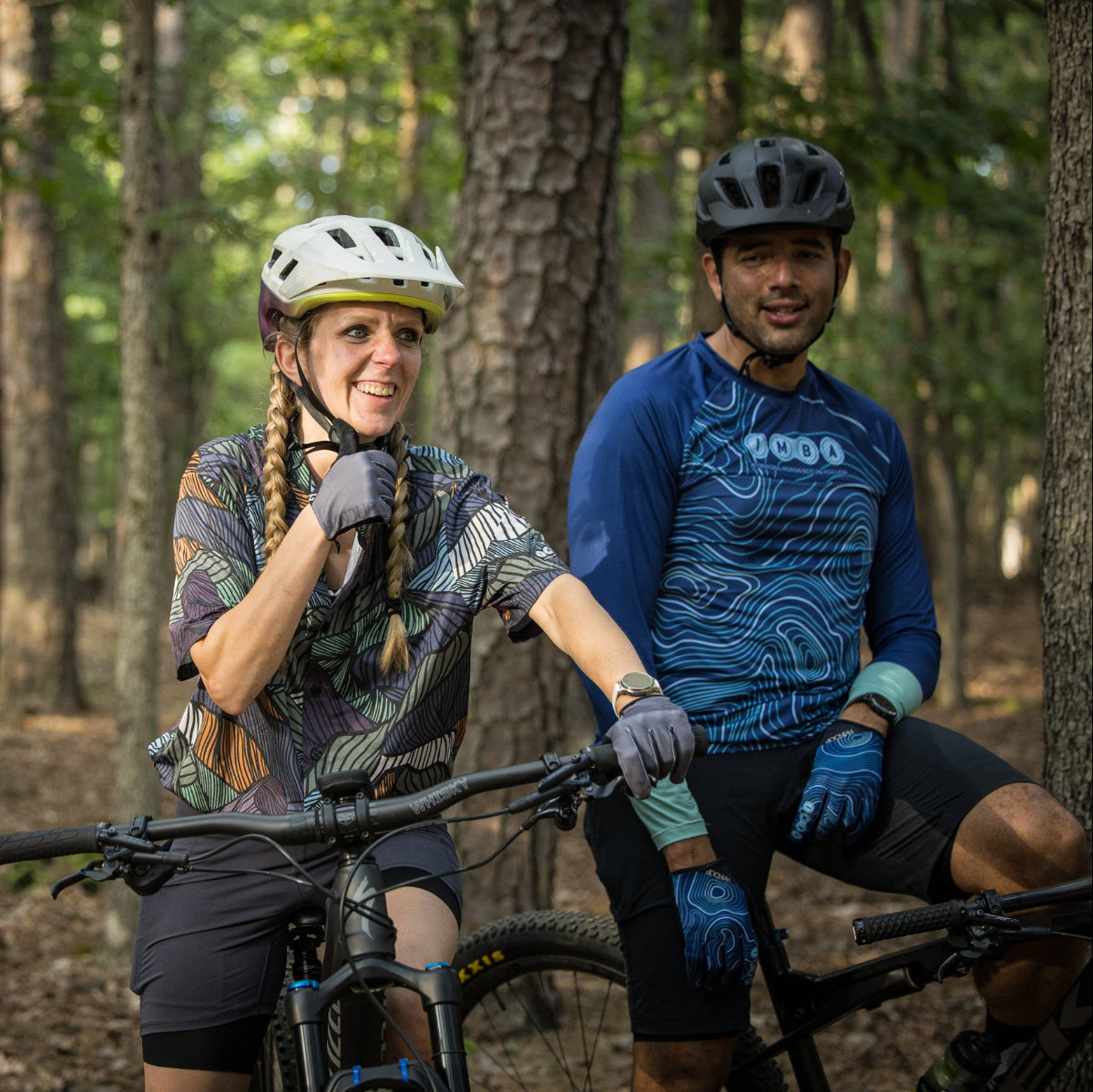Two mountain bikers in a forest setting, one of whom is adjusting their helmet.