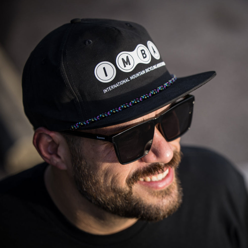 Man wearing a black cap with 'IMBO' logo and sunglasses, against a blurred background