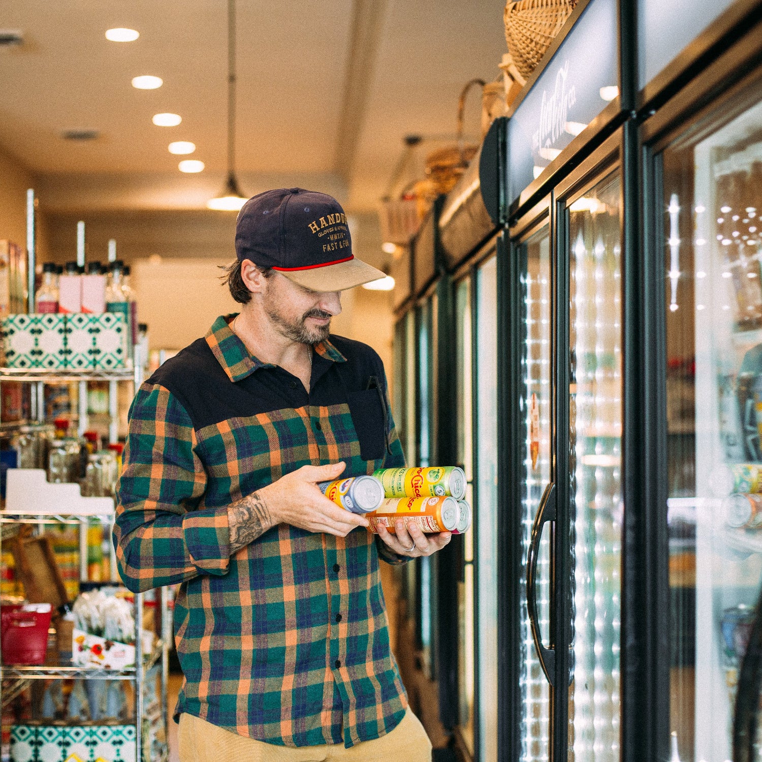man in a convenience store holding canned beverages and wearing a plaid flannel and hat