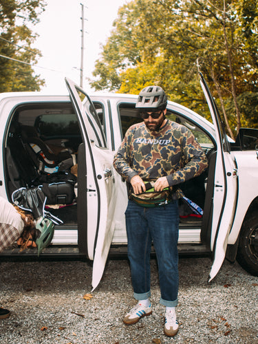Two people with a dog near an open white truck in a wooded area