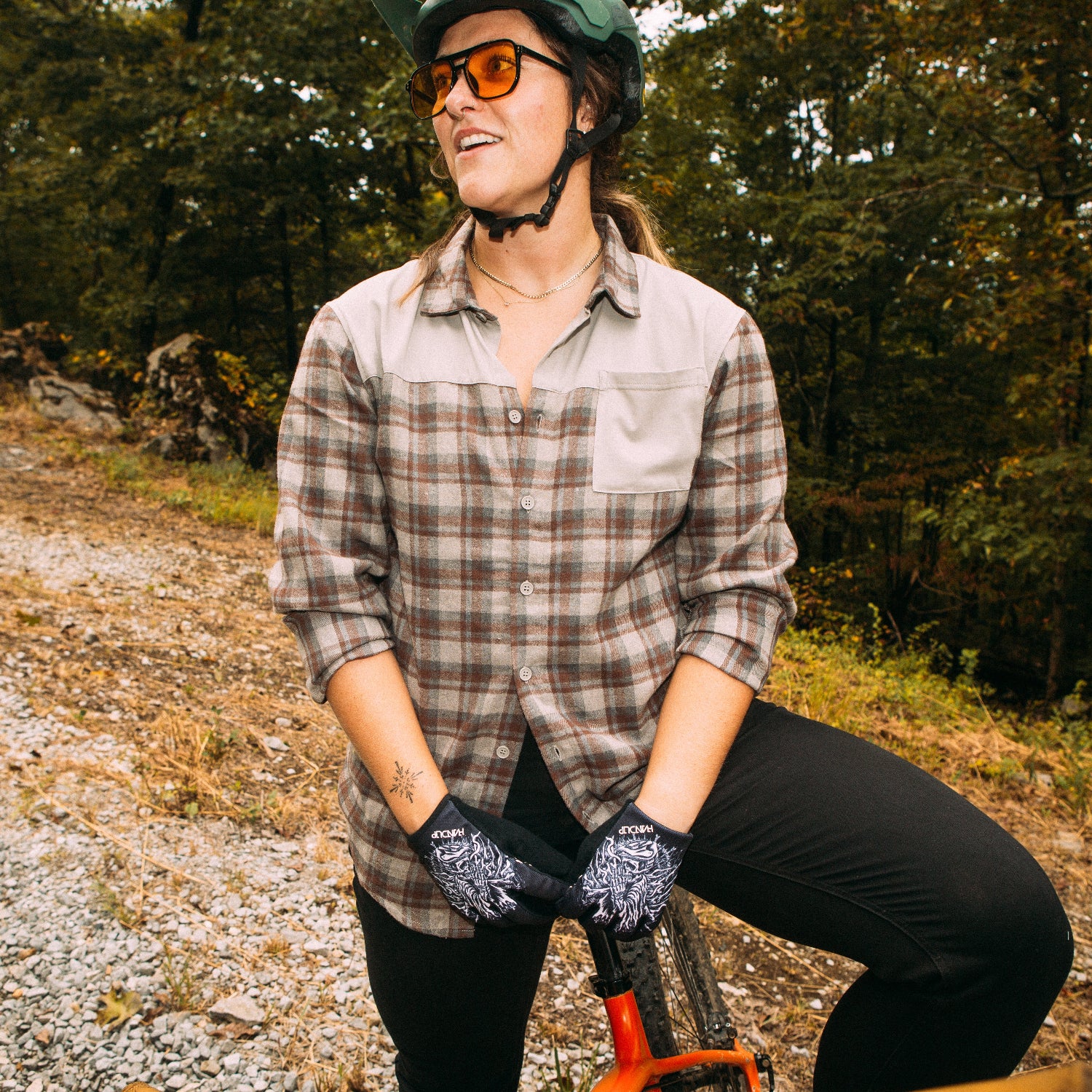 a woman wearing a flannel button up sitting on a mountain bike with trees and gravel in the background
