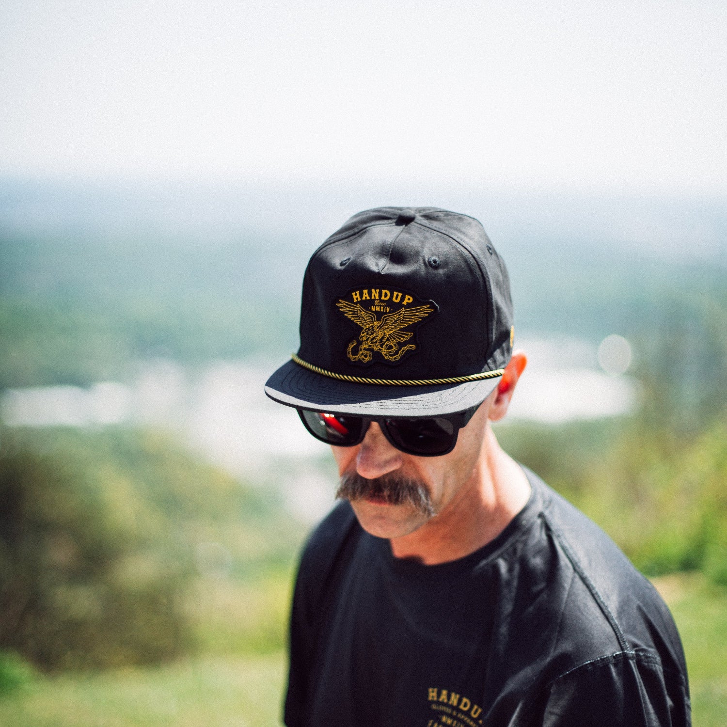 Man wearing a black cap with a logo and sunglasses, standing outdoors with a blurred natural background