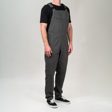 A man wearing dark gray bib overalls over a black t-shirt and black sneakers, standing against a light gray studio background.