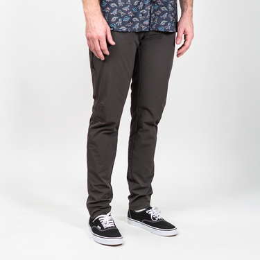 A person wearing slim-fit dark charcoal gray pants, paired with a patterned short-sleeve button-up shirt and black low-top sneakers, photographed against a clean light gray studio background.