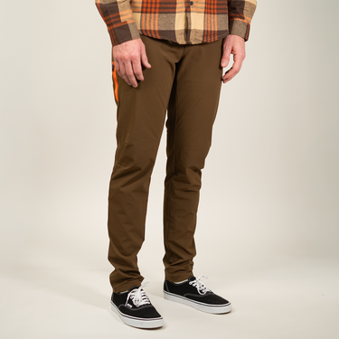 A person wearing slim-fit brown pants, a brown and orange plaid flannel shirt, and black low-top sneakers, photographed against a plain light beige studio background.