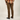 A person shown from the back wearing brown slim-fit pants, a brown and orange plaid flannel shirt, and black Vans sneakers against a light beige studio background.