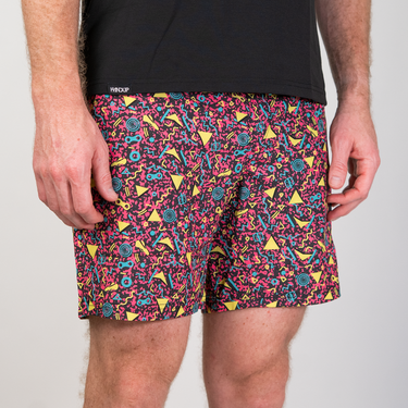 A close-up of a person wearing patterned athletic shorts and a black t-shirt against a light gray studio background. The shorts feature a dense multicolor abstract print with pink, yellow, blue, and black shapes.