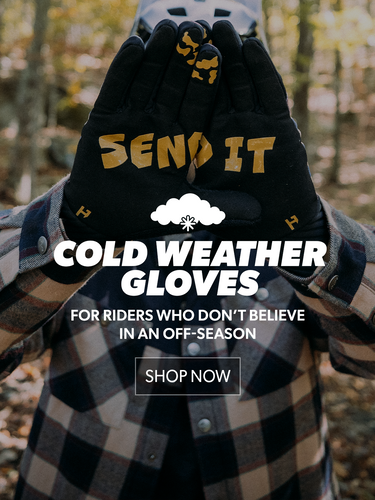 Person wearing black cold weather gloves with 'SEND IT' text, set against a blurred forest background.