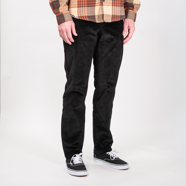 A person wearing black corduroy pants, a brown and orange plaid flannel shirt, and black canvas sneakers with white laces, standing against a light gray studio background.