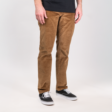 A person wearing brown corduroy pants, a black t-shirt, and black low-top sneakers with white laces and soles, standing against a light gray studio background.