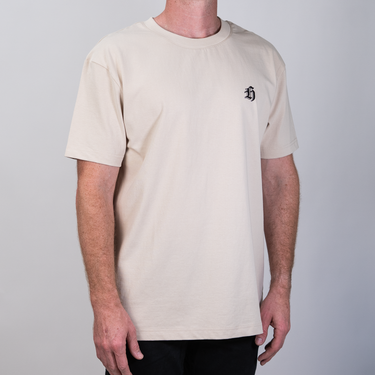 A man wearing a light beige crewneck t-shirt with a small black embroidered logo on the left chest, photographed against a neutral gray background.