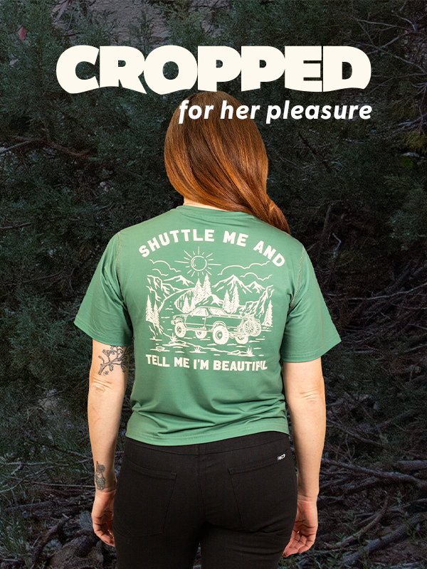 Person wearing a green t-shirt with text and graphics, standing against a dark forest background.