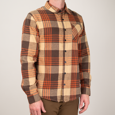 A man wearing a long-sleeve button-up flannel shirt with a brown, beige, and orange plaid pattern, paired with brown pants, photographed against a solid light gray background.