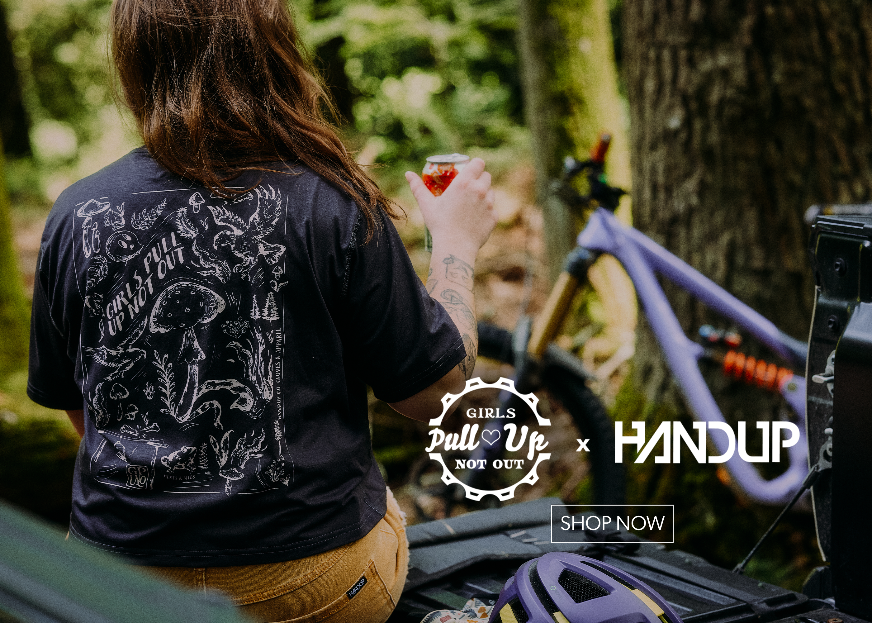 A woman seen from behind wearing a black graphic t-shirt with white mushroom and forest illustrations, sitting outdoors next to a purple mountain bike and a trunk. The image features 'Girls Pull-Up Not Out' and 'HANDUP' branding with a 'SHOP NOW' button overlay.