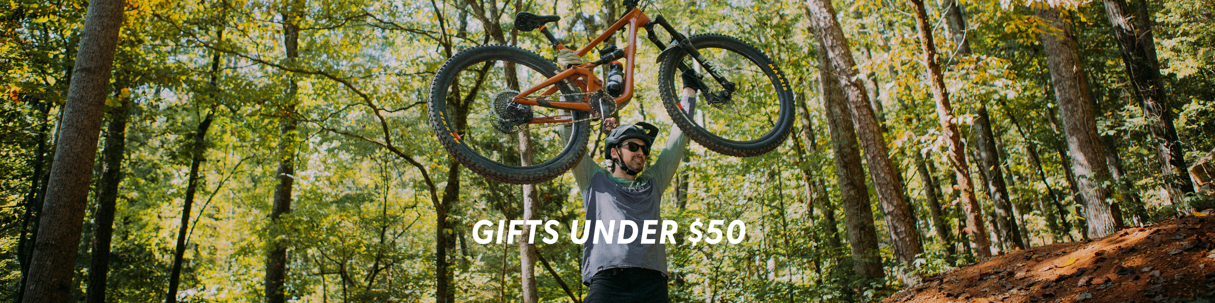 Man holding a mountain bike in a forest with 'Gifts Under $50' text overlay.