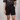 Person wearing black shorts and a black t-shirt with a tie, on a plain background