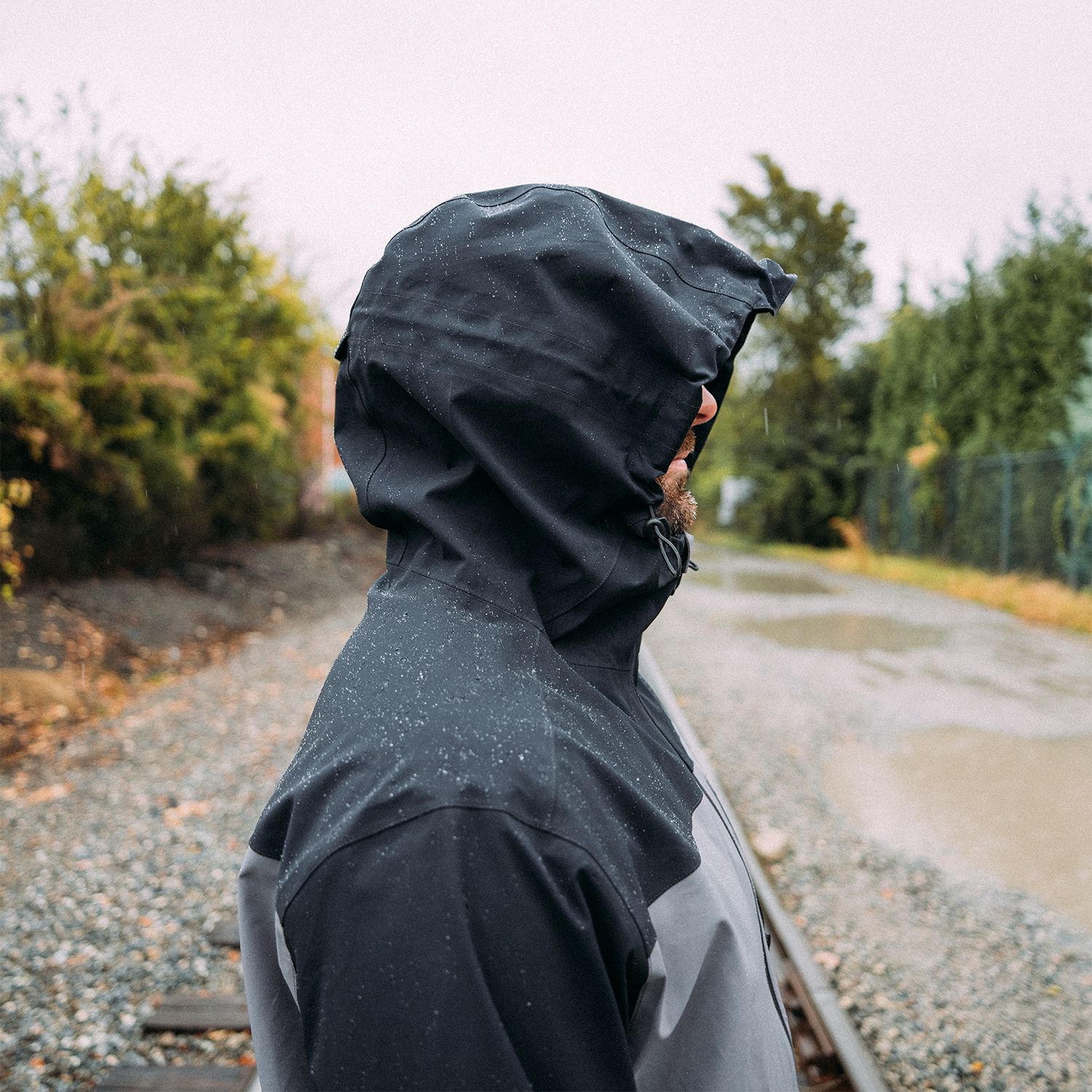 Person wearing a black raincoat on a rainy day with trees and a road in the background.