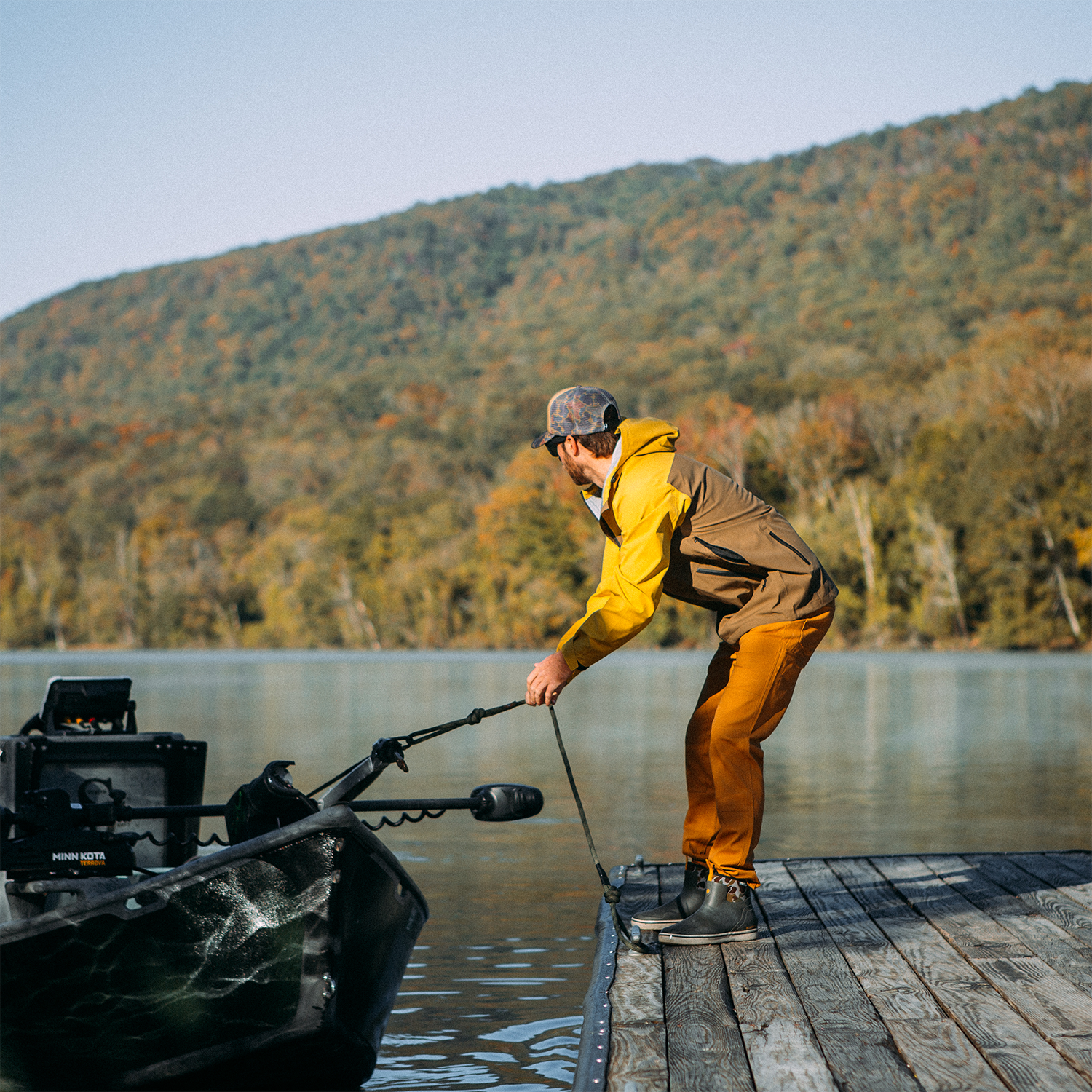Man in yellow jacket and orange pants standing on a dock with a boat, surrounded by a scenic lake and mountains.