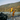 Man in yellow jacket and orange pants standing on a dock with a boat, surrounded by a scenic lake and mountains.