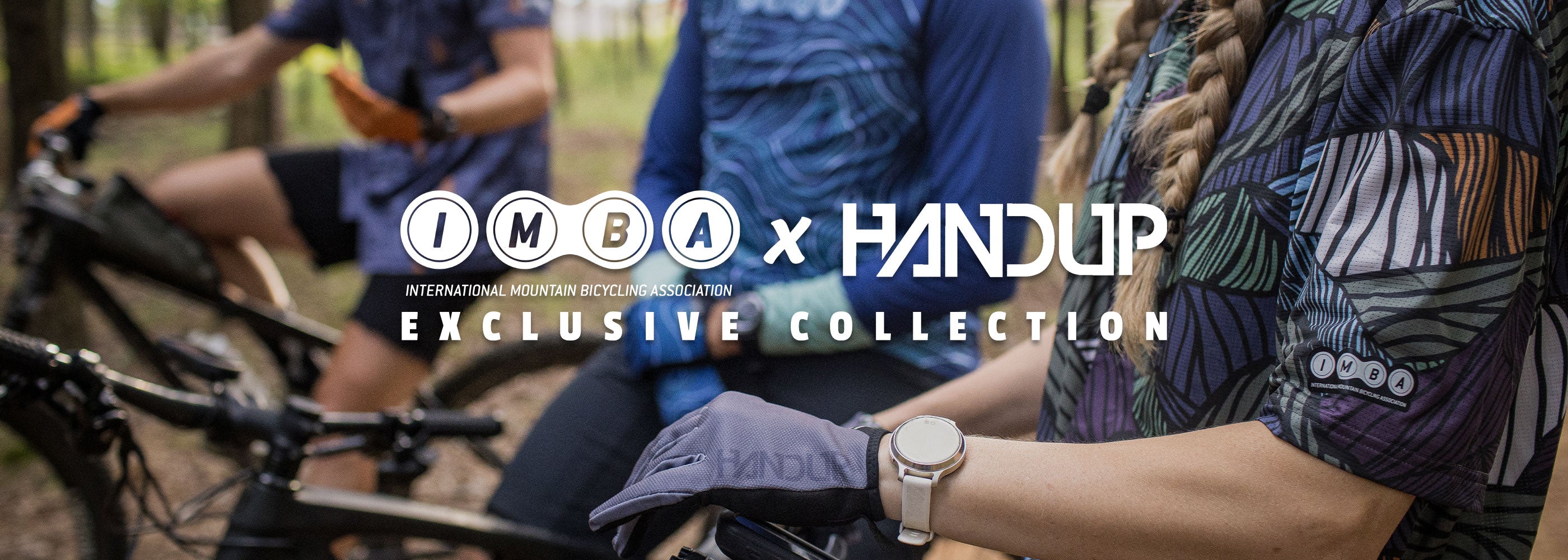Two people with bicycles in a forest setting, featuring the IMBA x Handup Exclusive Collection logo.