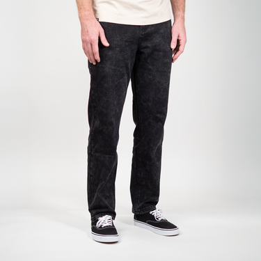 A person wearing black acid-wash straight-leg jeans, a cream t-shirt, and black canvas sneakers with white laces, standing against a light gray studio background.