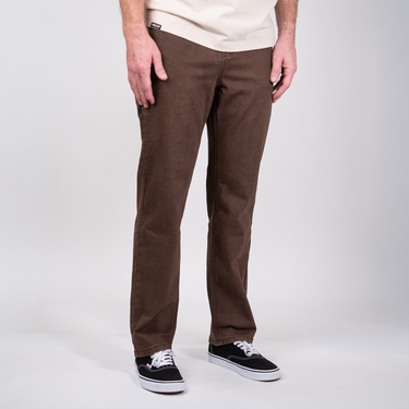 A person wearing brown straight-leg corduroy pants, a light beige t-shirt, and black canvas sneakers with white soles, standing against a light gray studio background.