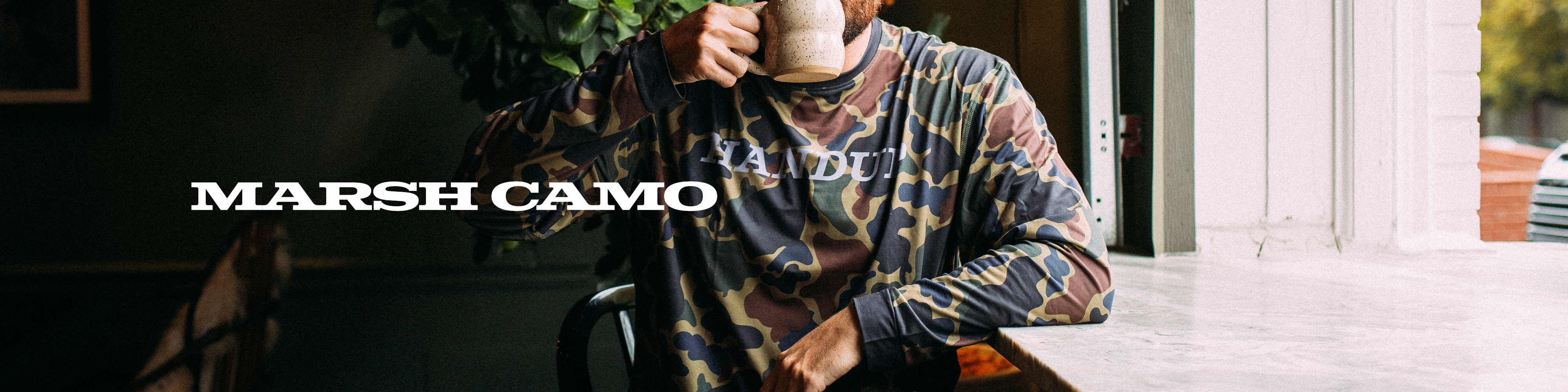 Person wearing a patterned shirt with 'Marsh Camo' branding, holding a coffee cup indoors.