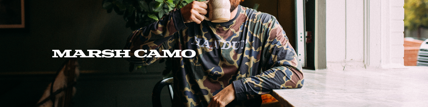 Person wearing a patterned shirt with 'Marsh Camo' branding, holding a coffee cup indoors.