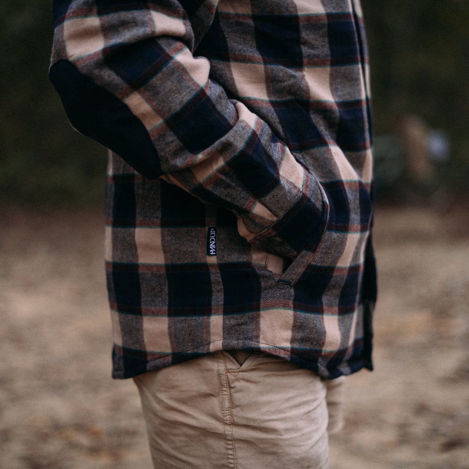 person wearing a plaid flannel jacket and khaki pants outdoors with a blurred background.