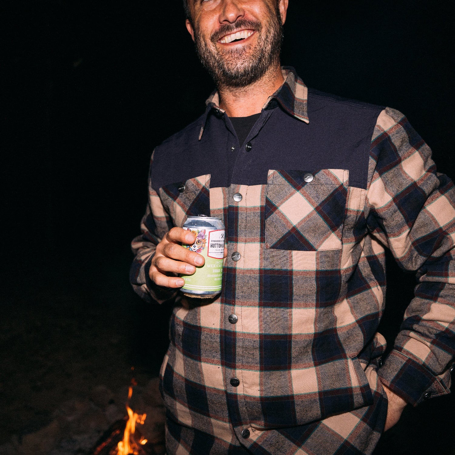 Man in plaid shirt holding a can at night with a fire in the background