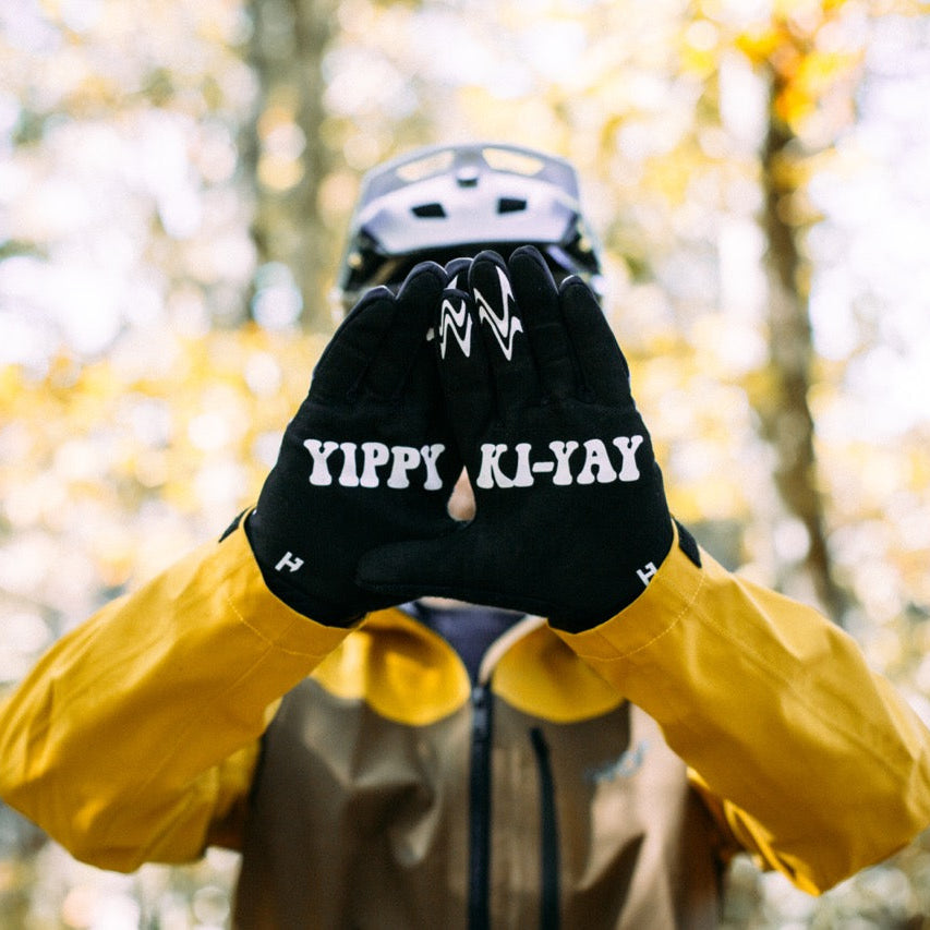 Person wearing a black glove with 'YIPPIE KI YAY' text, yellow jacket, and helmet in a forest setting.