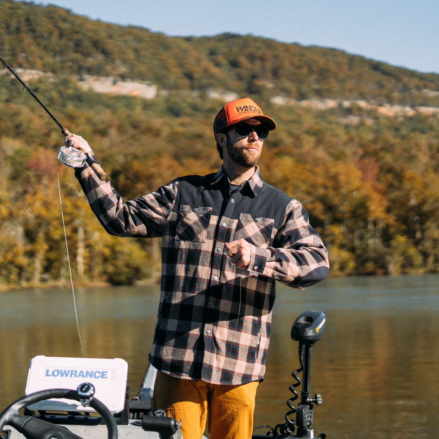 a man wearing a flannel plaid jacket and orange hat, holding a fishing pole with water and trees in the background