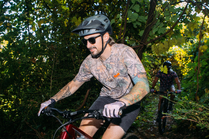 Man riding a mountain bike through a forest with another cyclist in the background.