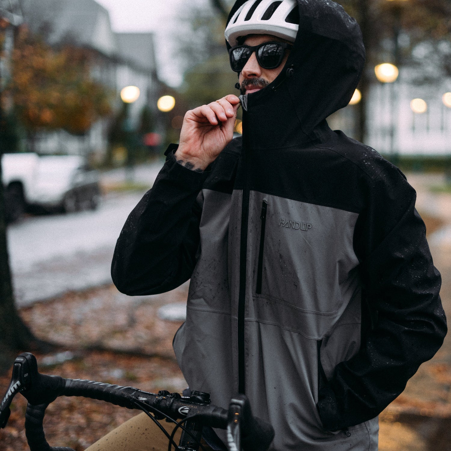 Person wearing a black and gray raincoat with a helmet and sunglasses, standing outdoors on a rainy day.