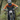 A full-face mountain biker wearing an orange and navy long-sleeve jersey, black pants, and a black helmet with yellow goggles rides a black full-suspension mountain bike through a forest.
