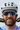 A smiling cyclist wearing a white helmet, mirrored sunglasses, and a blue and pink tie-dye jersey, holding up black gloves with colorful finger sleeves. The gloves feature gold text that reads 'STAY THRIFT' and 'THANK GRAVEL'.