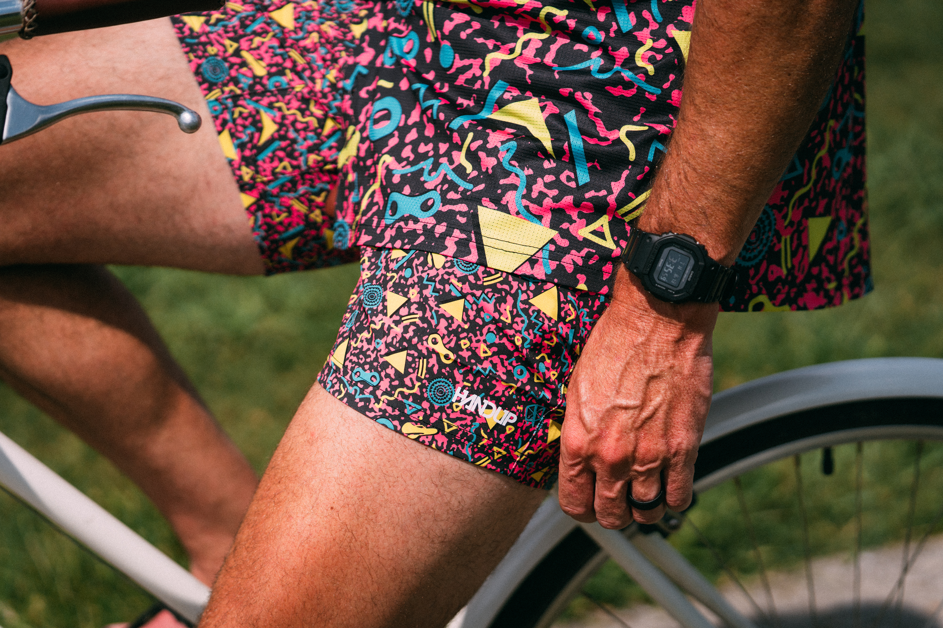 Close-up of a person wearing colorful geometric patterned cycling shorts while sitting on a bicycle outdoors. The shorts feature a busy abstract print in pink, yellow, blue, and black, with a matching compression-style thigh band.